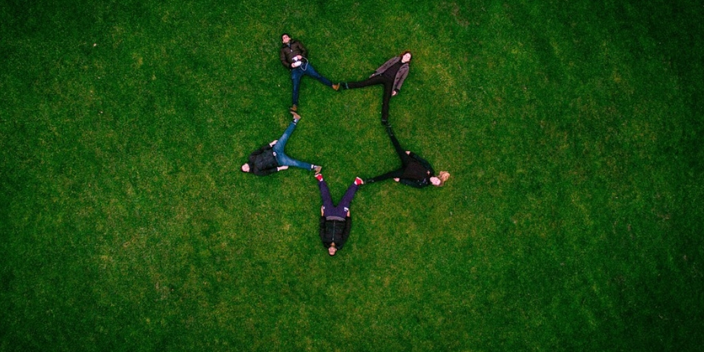 Image with five people resting on the ground with grass. There feet are joined to each other in form of a star.