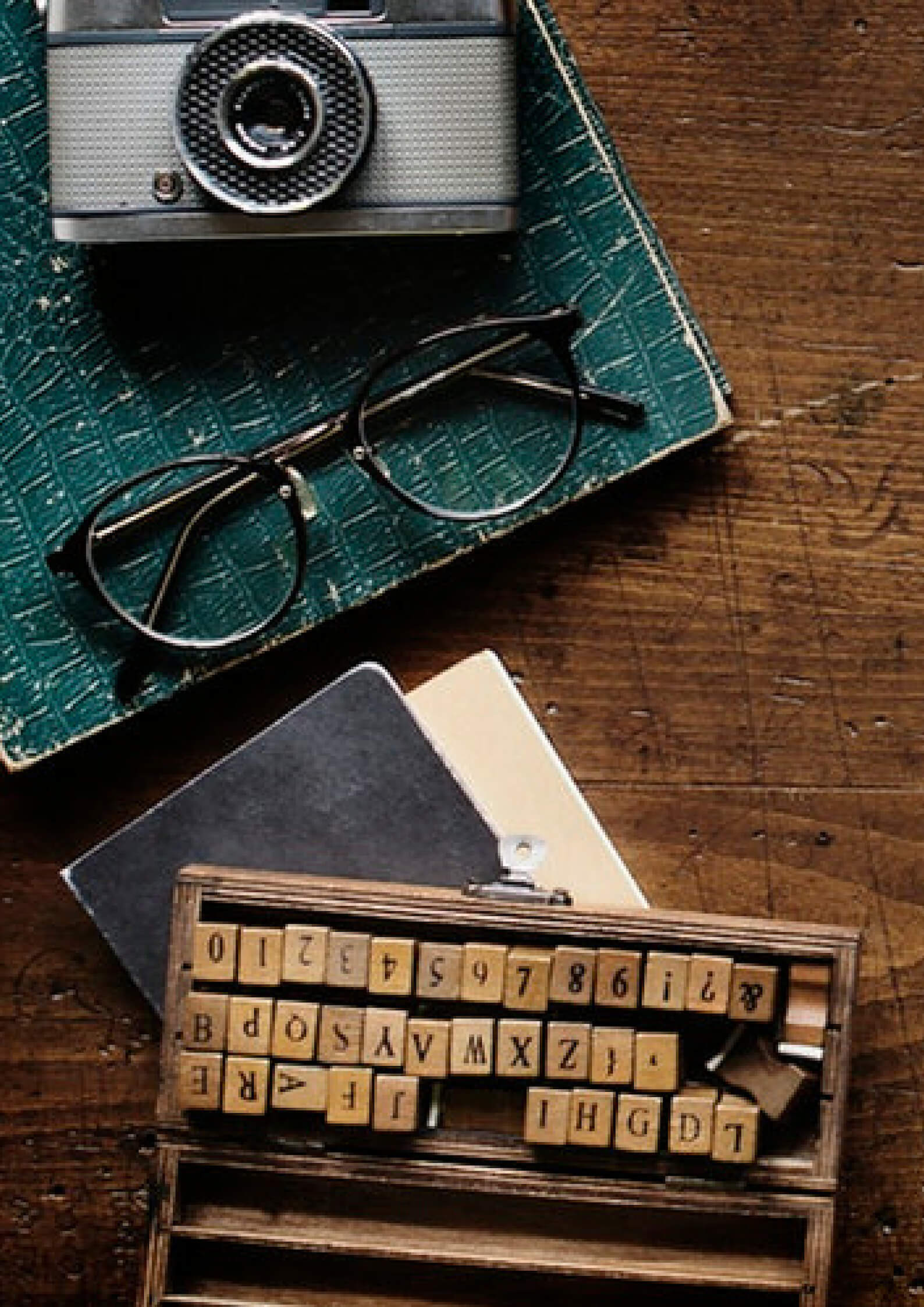 Image of a green notebook on which a camera and glasses are placed with a word constructor games on a wooden background. 
