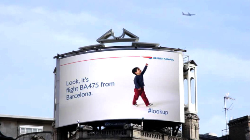 A big screen shows a child pointing his fingers upwards while a plane flies in the sky