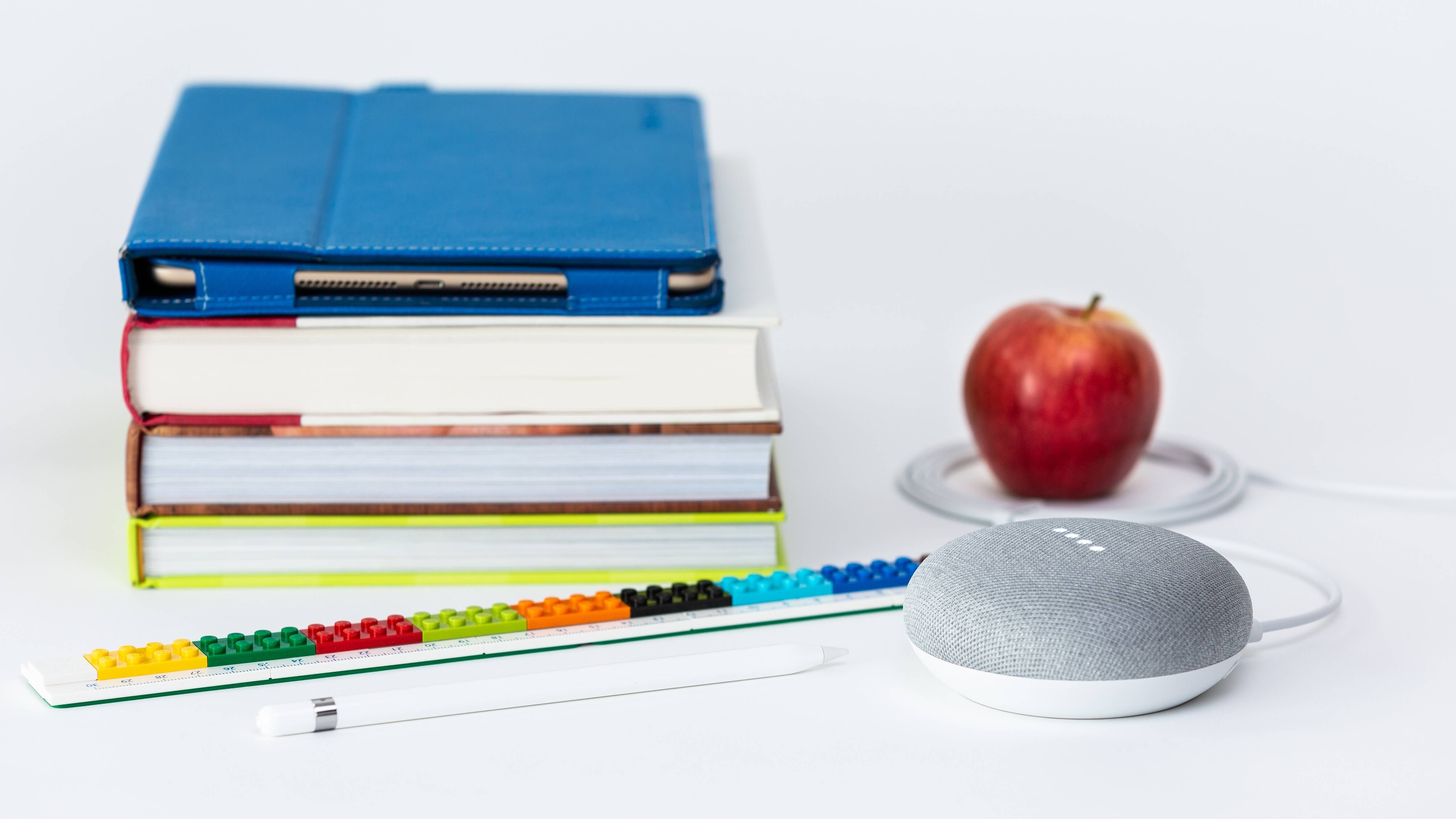 A pile of books, a voice interface device, a pen and an apple kept close to each other