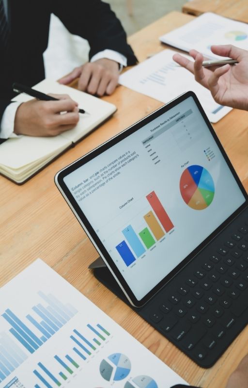 laptop displaying pie chart and bar graph kept on a brown desk and two hands writing on a notebook kept on the same desk