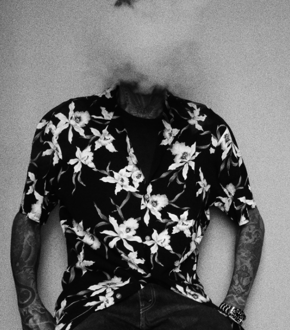 A black and white image of a man wearing a shirt with flower designs and his face blurred by smoke