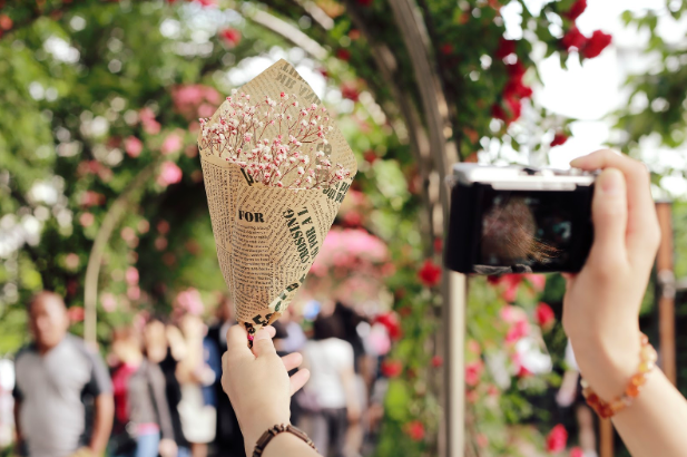 Image, representing microservices, showing hands of a person clicking the picture of a handmade bouquet in one and camera on the other and blurred flowers, trees, people in the background