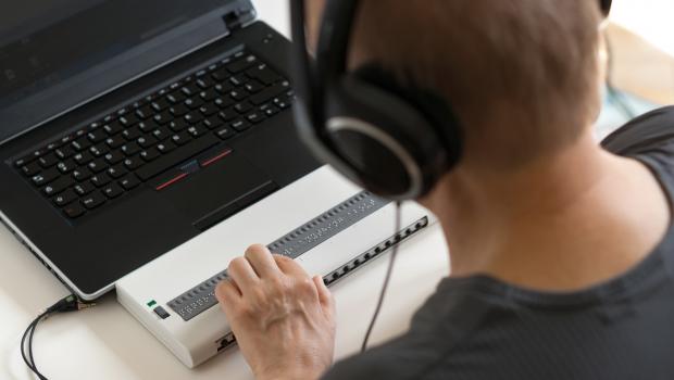 A person with headphones, touching the braille tool connected to a black laptop