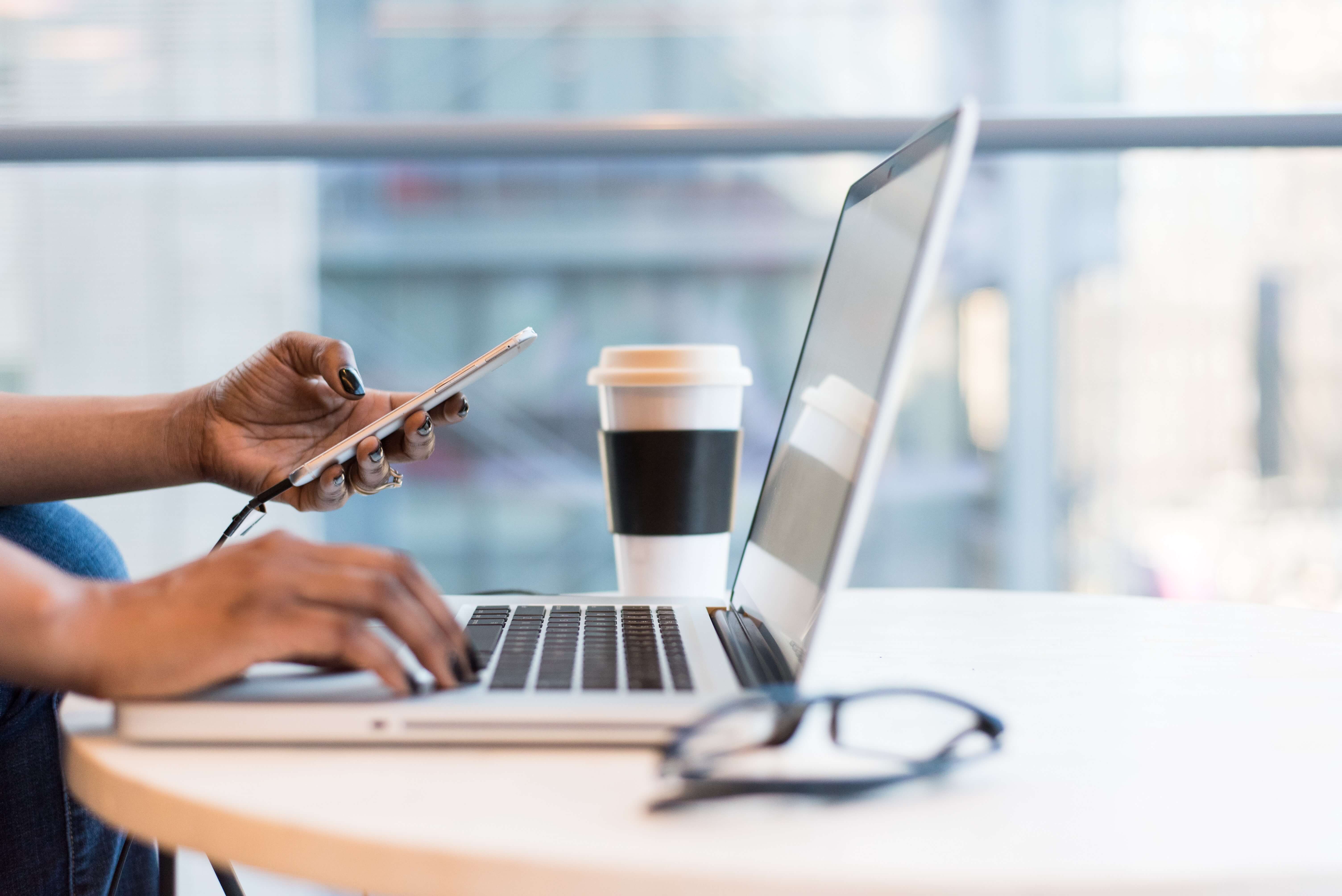 a laptop and coffee cup on the table while a woman uses her phone