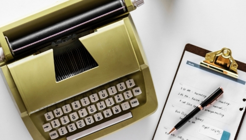 Gold typewriter on left and a sheet of paper on a pad with pen on right