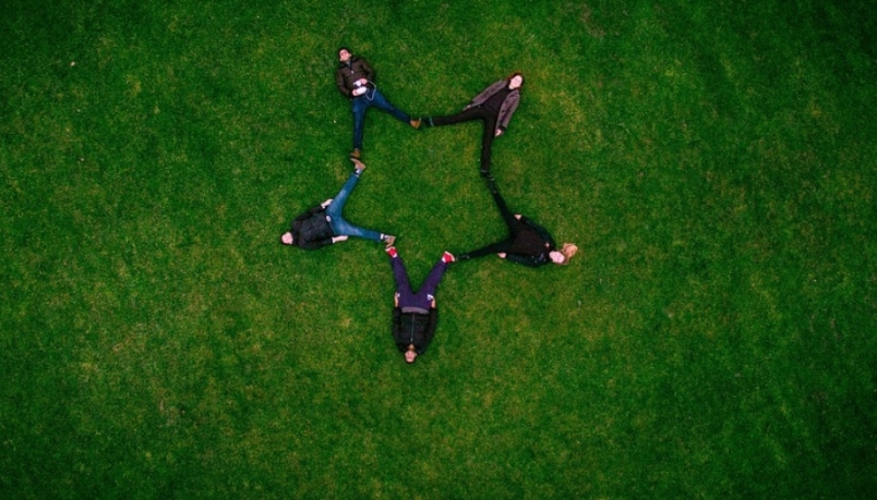 Image with five people resting on the ground with grass. There feet are joined to each other in form of a star.