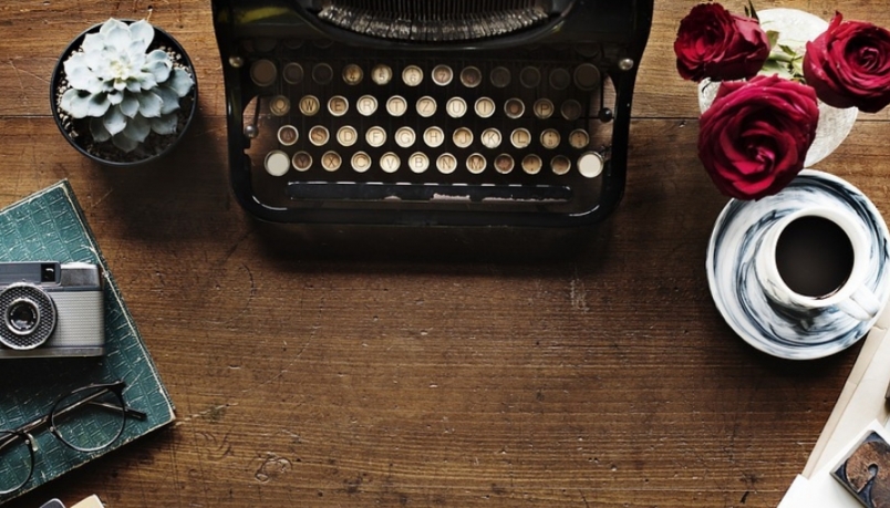 Image of a typewriter placed at the center of a wooden background where there is a tea pot and a flower vase on the left side and a camera on right side 