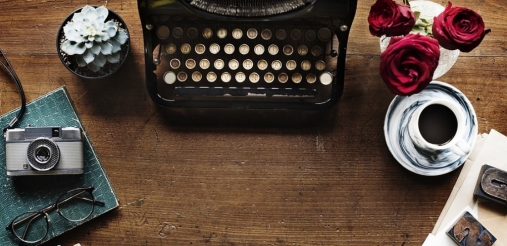 Image of a typewriter placed at the center of a wooden background where there is a tea pot and a flower vase on the left side and a camera on right side
