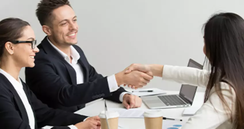 3 people are siting, 2 of them are hand shacking and the other person is smiling