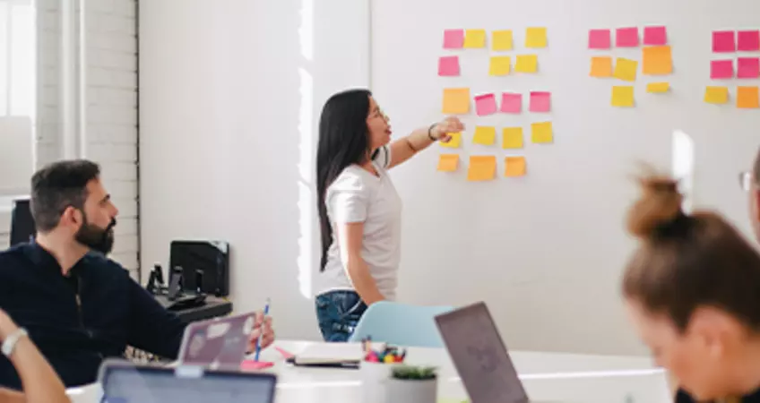 a women is explaining something on a board to a group of men and women siting with laptops